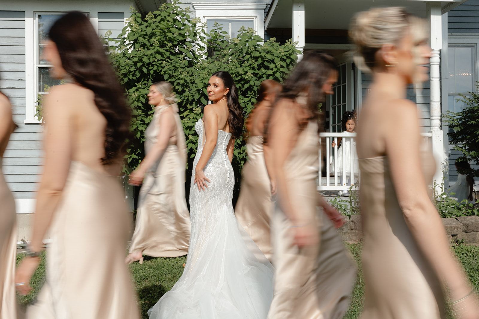Bride surrounded by bridesmaids in champagne satin walking past a porch