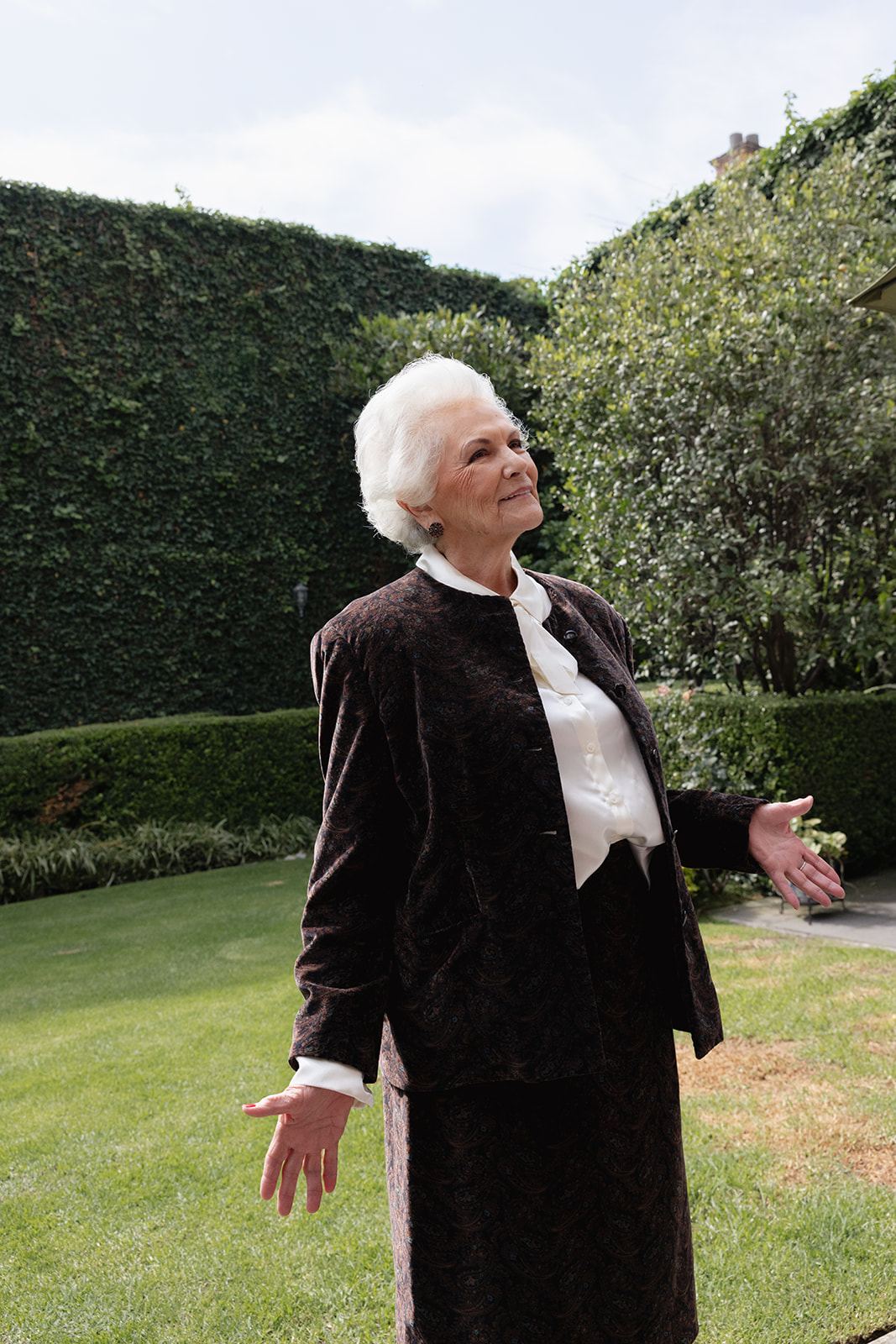 Elderly woman in velvet smiling in a garden with tall hedges