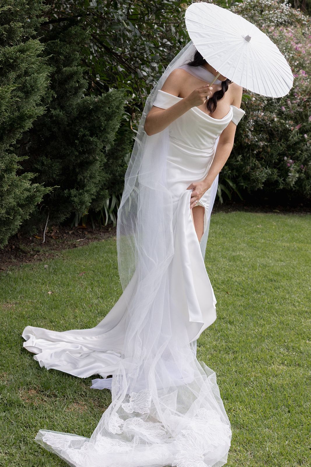 Bride under a white parasol adjusting her off-shoulder gown on the grass